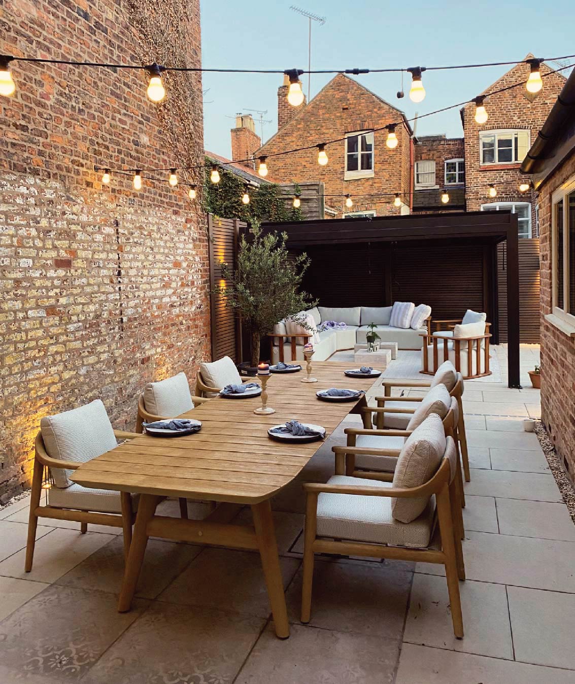 A brick-walled courtyard with a large N1 teak dining table, cushioned chairs, outdoor seating, and string lights overhead.