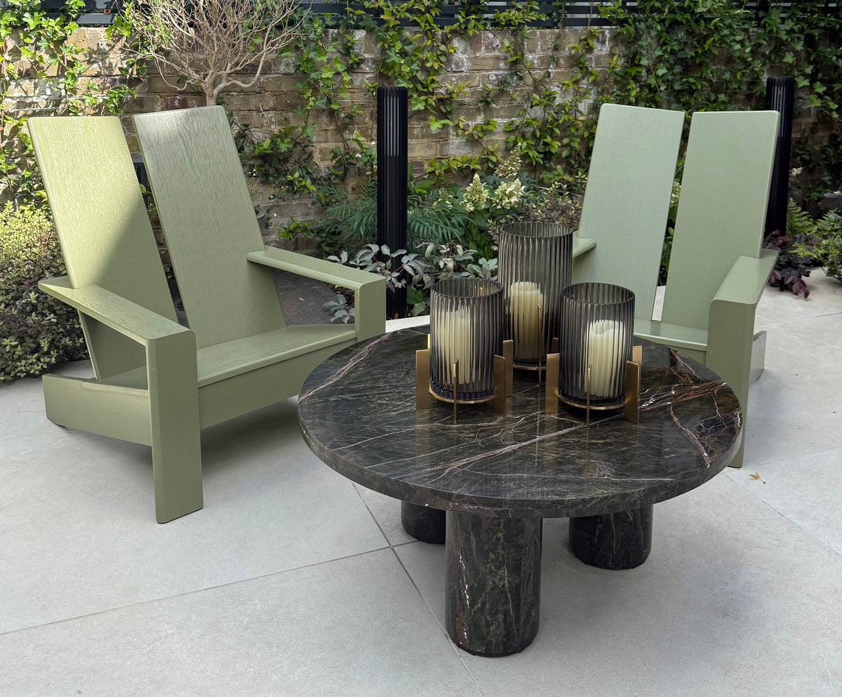 A modern outdoor patio furniture set featuring two sage green Adirondack-style chairs and a round dark marble coffee table with three ribbed glass candle holders, set against a stone wall with ivy.