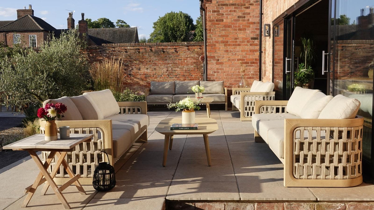 Outdoor patio furniture set with woven wicker-style frames, light beige cushions, and matching coffee tables arranged on a paved terrace beside a brick wall and glass doors.