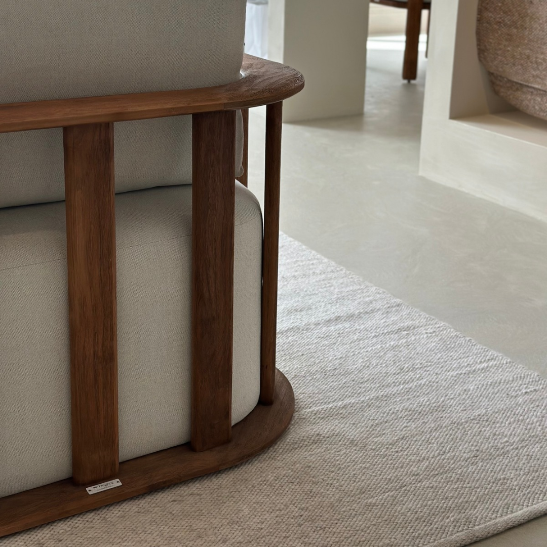 Close-up of a modern armchair with light beige upholstered cushions and a curved dark brown wooden frame, placed on a textured light beige rug in a minimalist interior setting.