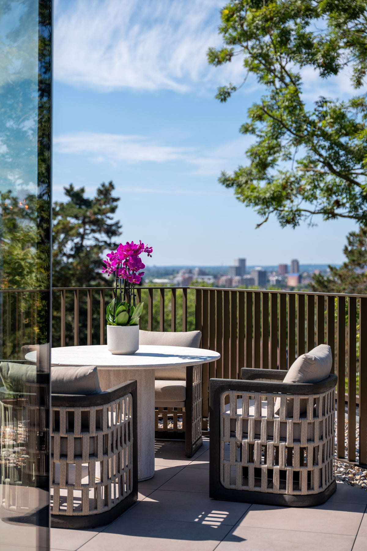 Modern outdoor patio furniture set with a round white table and two woven gray armchairs on a tiled terrace, featuring a vibrant purple orchid in a white pot and a scenic city skyline in the background under a blue sky.
